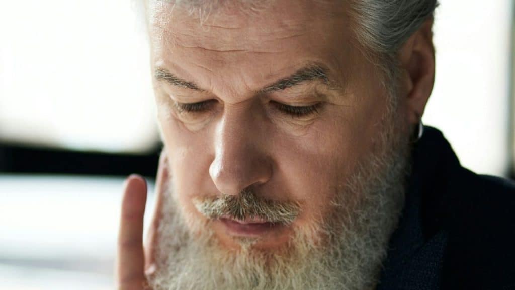 A close-up of an older man with a white beard and hair looking down intently, holding his hand near his face.