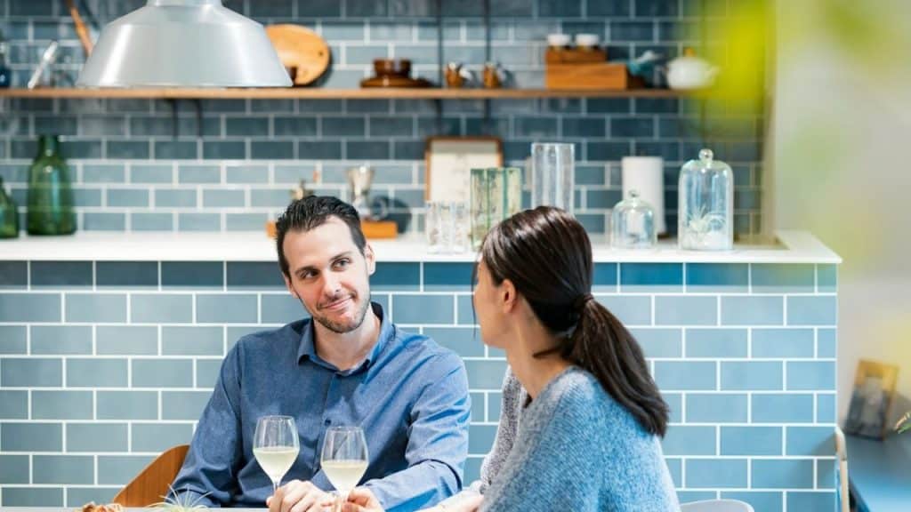 A man and woman sit across from each other at a kitchen table, looking at each other over wine glasses.