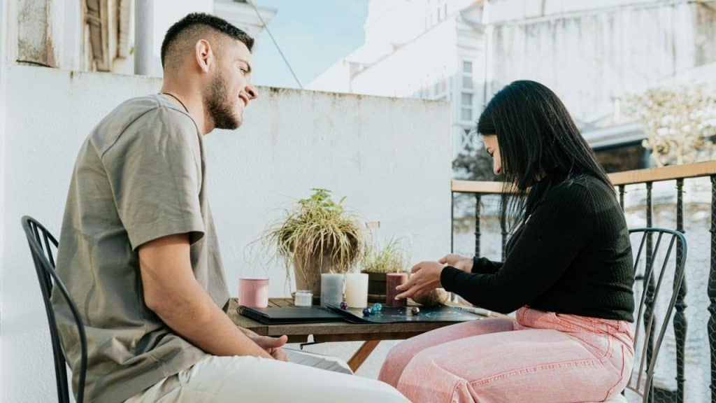 A smiling man and a woman are sitting across from each other at a table on a balcony.