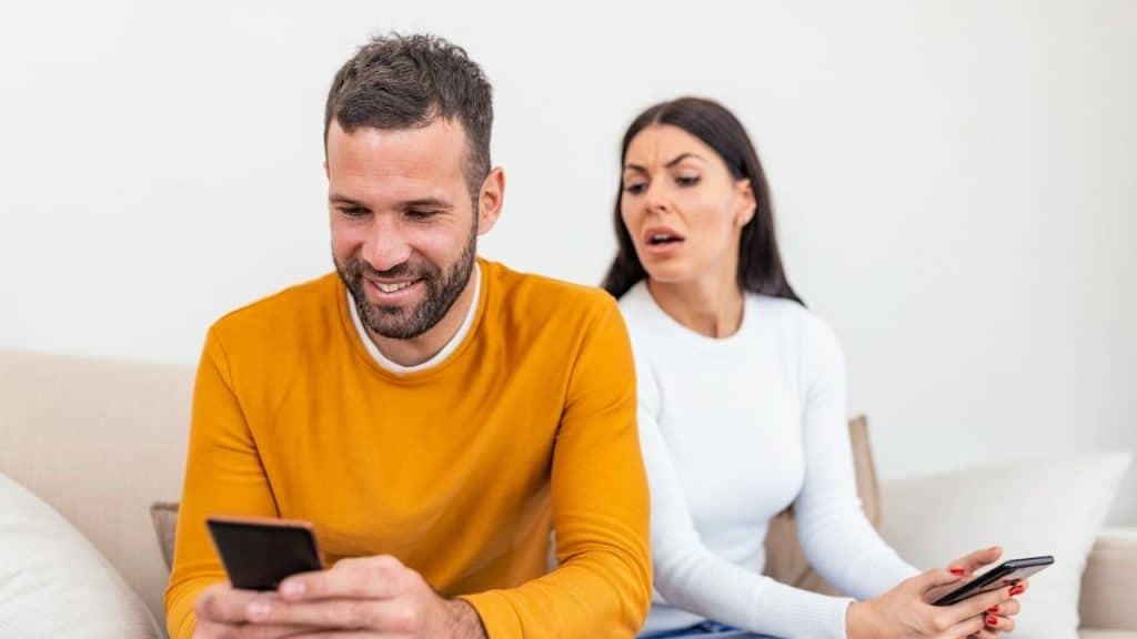 A smiling man checks his phone while a woman looks over his shoulder suspiciously.