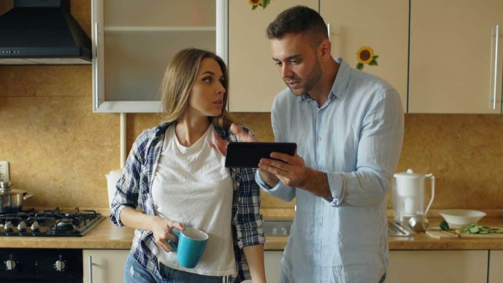 A man shows a digital tablet to a woman in a plaid shirt in a kitchen.