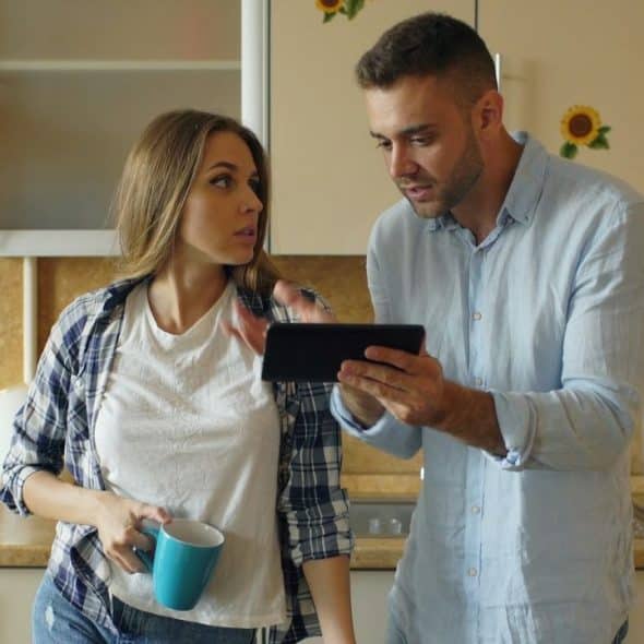 A man shows a digital tablet to a woman in a plaid shirt in a kitchen.