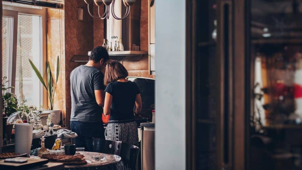 A couple stands close together, cooking in a warmly lit kitchen with wooden cabinets.