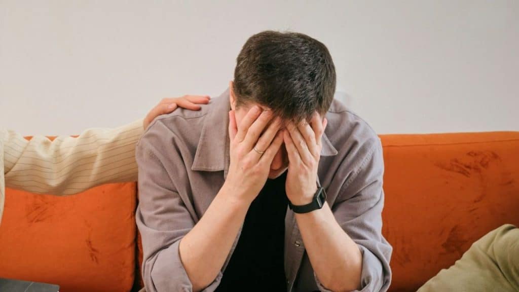 Man seated on an orange couch with his face in his hands, being comforted by a hand on his shoulder.