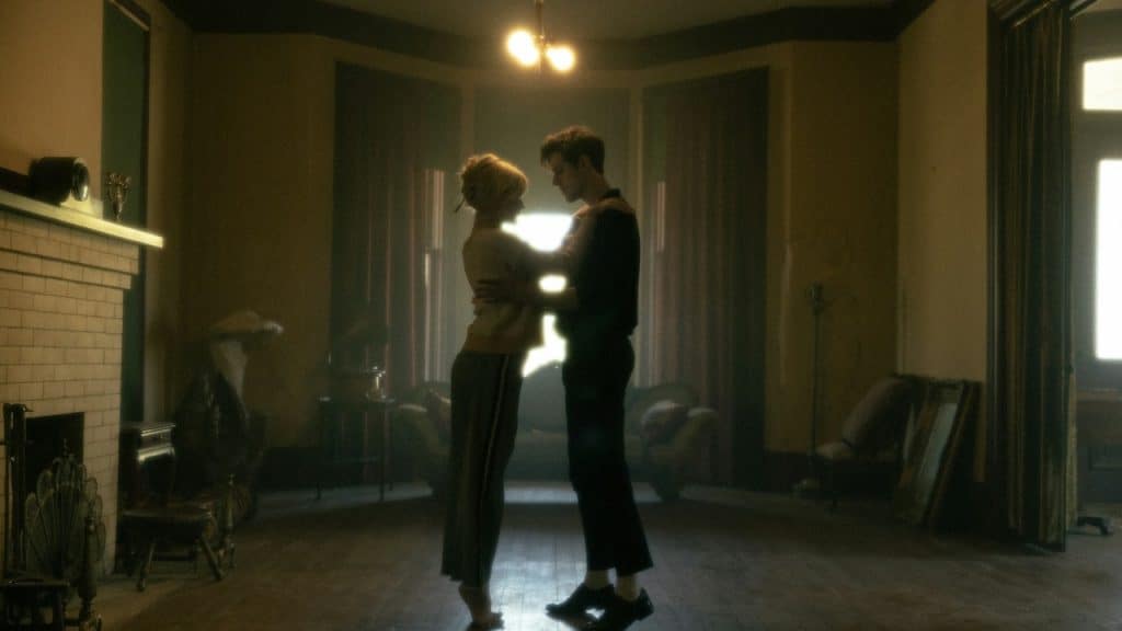 A man and a woman stand facing each other in a dimly lit, large room with a wooden floor, a fireplace, and tall windows.