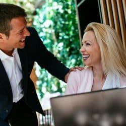 A smiling man with his hand on a smiling woman's shoulder at an outdoor cafe table with a laptop.