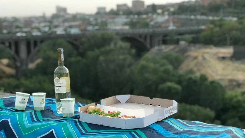 A partially eaten pizza in a box, a bottle of white wine, and three cups sit on a blanket overlooking a cityscape and a bridge.