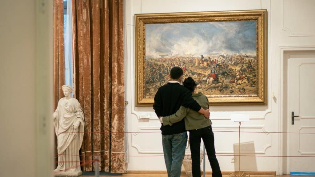 A couple stands in an art museum, facing a large historical painting of a battle, with their arms around each other.