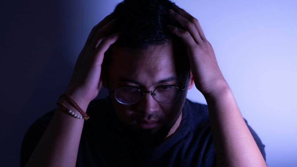 Man with glasses and a beard holding his head in his hands in a dimly lit space.