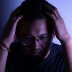 Man with glasses and a beard holding his head in his hands in a dimly lit space.