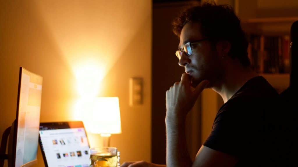 Man with glasses and hand on chin working on a laptop at night with a lamp.