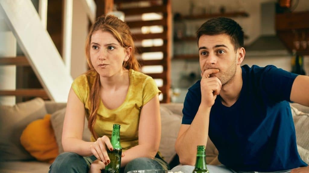 Two young adults sitting on a couch, holding beer bottles and watching something intently.