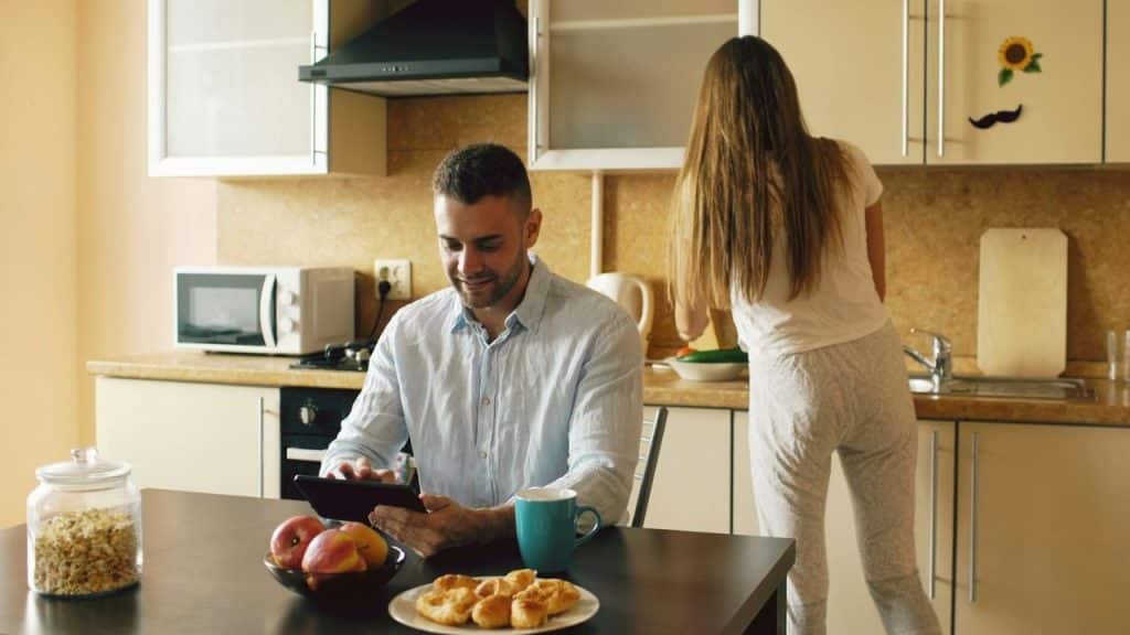 Man at a table looking at a tablet while a woman works in the kitchen background.