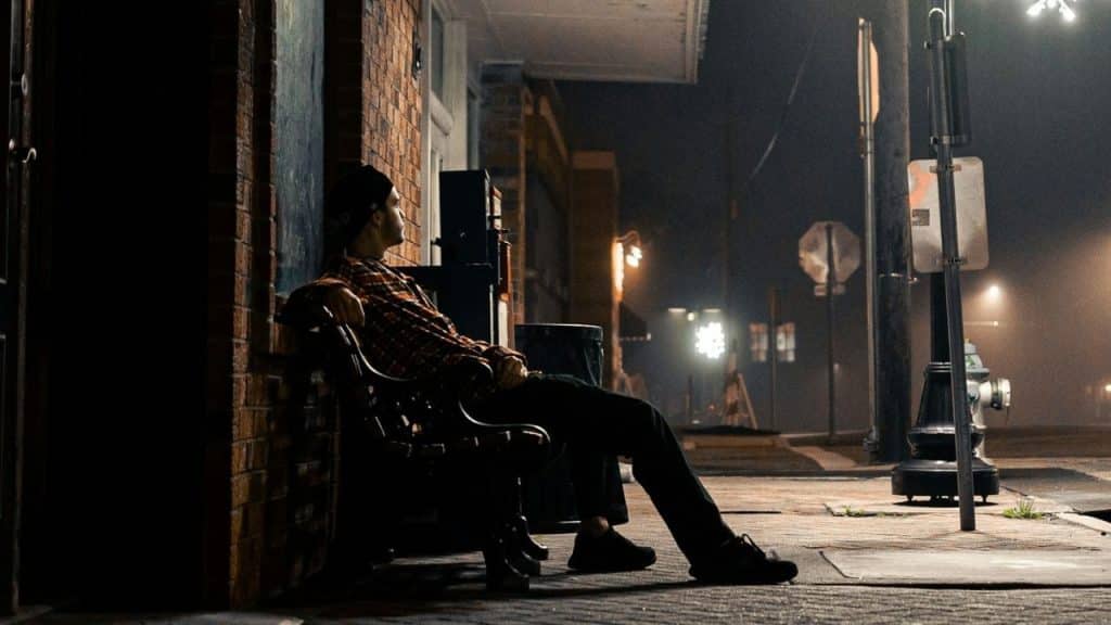 Man in a flannel shirt and beanie sitting alone on a bench outside at night.