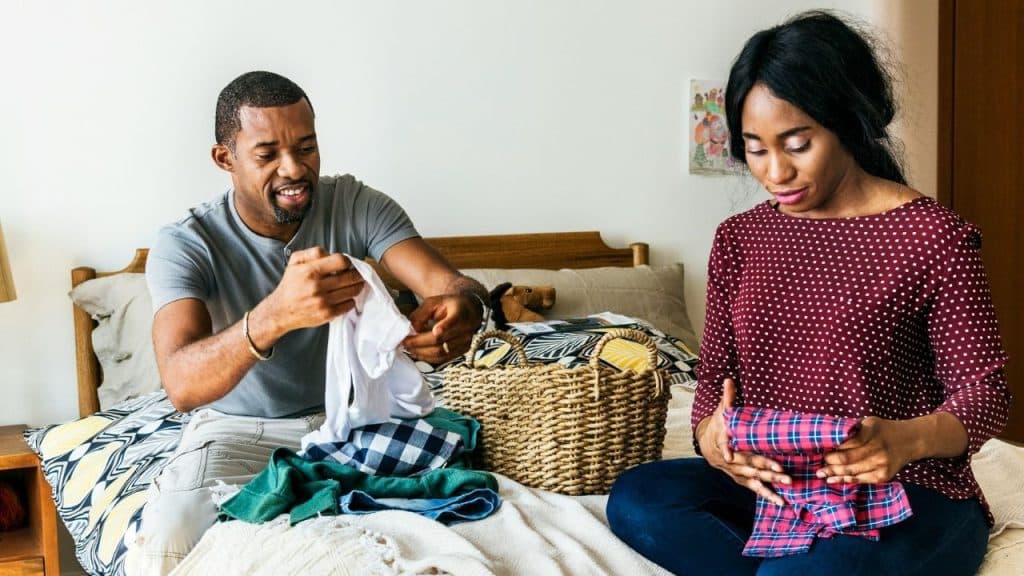 Man and woman sitting on a bed folding laundry from a wicker basket.