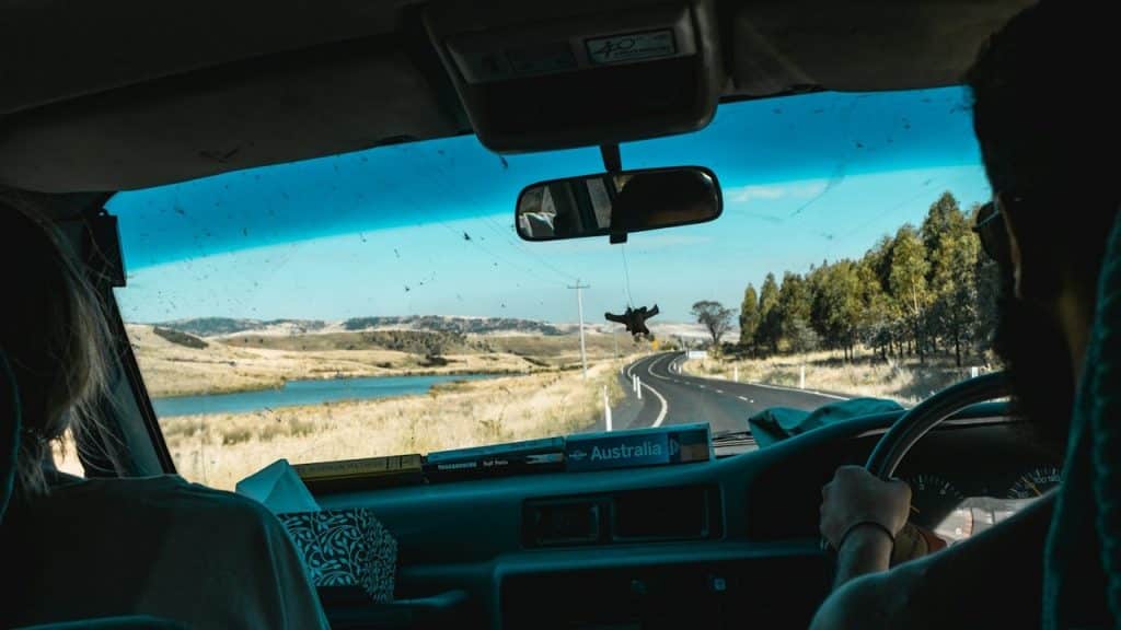 The view from inside a car as a person drives along a winding road through grassy hills and past a body of water under a bright blue sky.