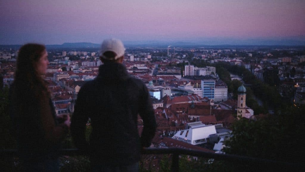 Two people stand with their backs to the camera, looking out over a city skyline.