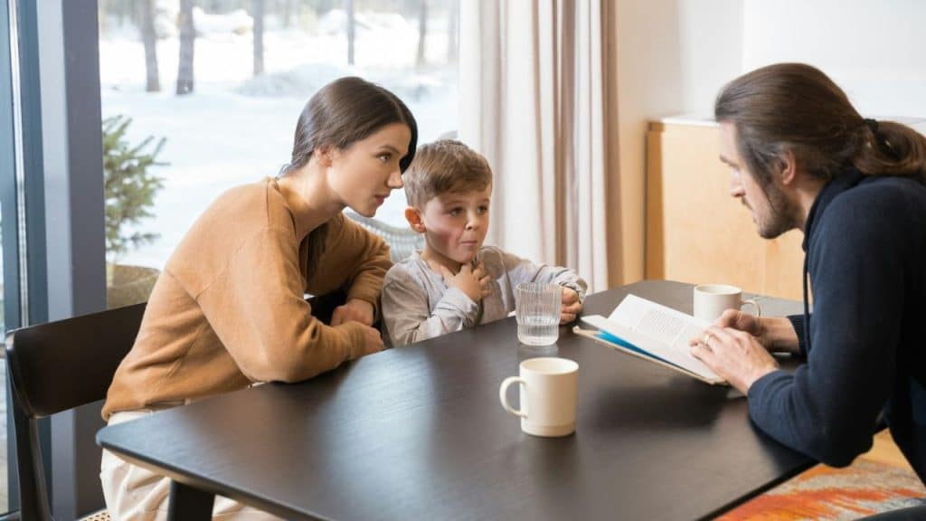 Woman and boy listening as a man reads a book at a table with mugs.
