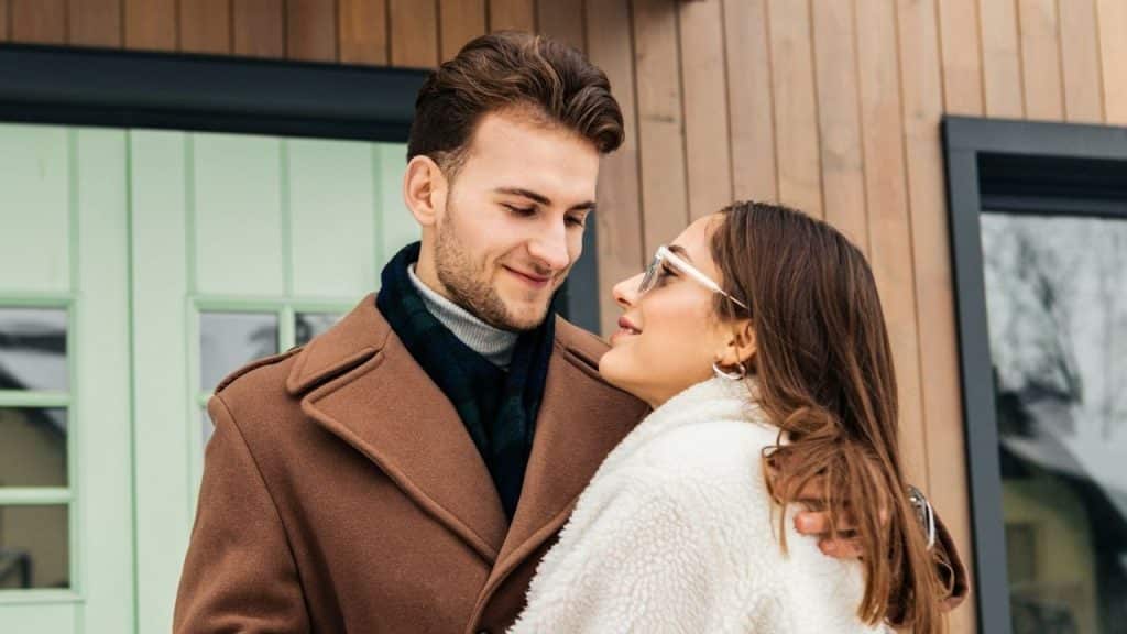 Couple warmly embracing and smiling at each other in front of a modern wooden house.