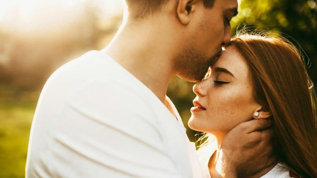 Man in a white shirt kissing a woman's forehead in a sunlit outdoor embrace.