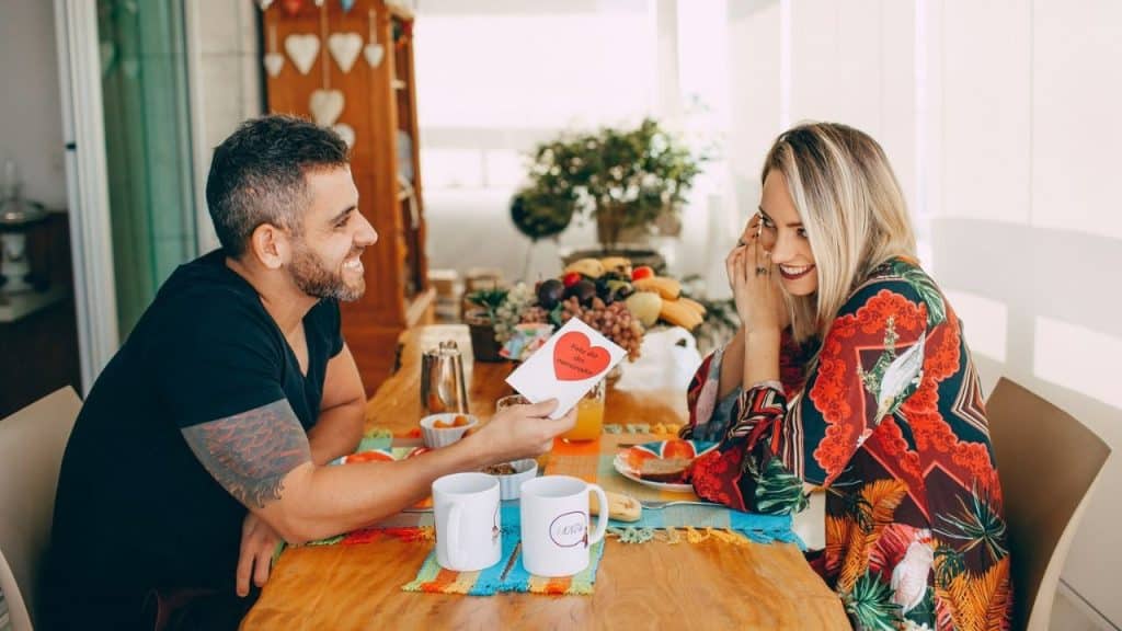 Smiling man handing a woman a card with a red heart at a breakfast table.
