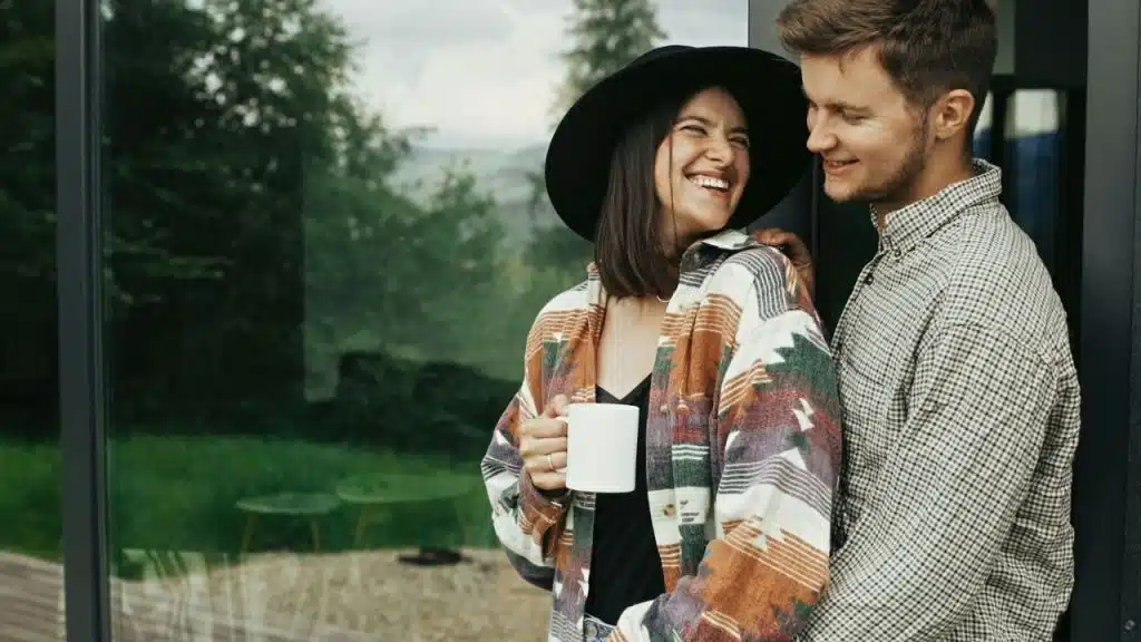 Couple standing by a glass door, woman in a hat laughing and holding a white mug.