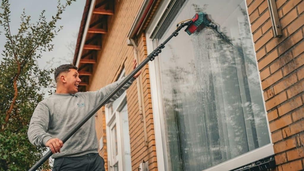 Man in a grey sweatshirt smiling while using a long-handled brush to clean a house window.