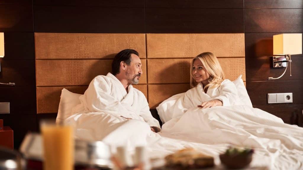A smiling couple in white bathrobes is having breakfast in bed, looking at each other in a luxurious hotel room.