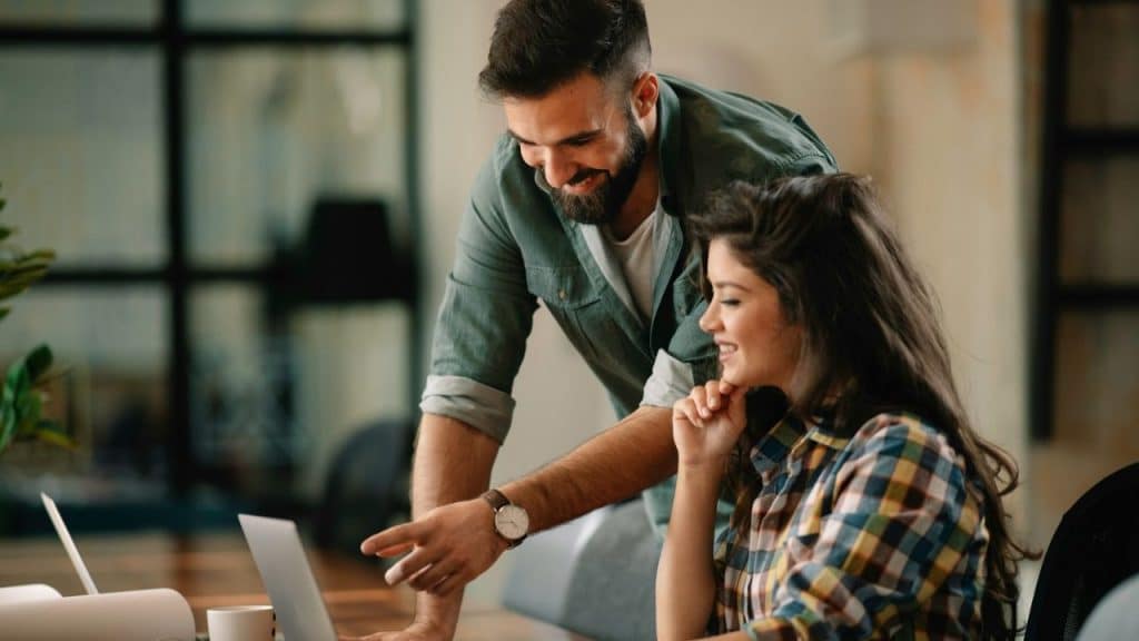 Man with a beard and a woman smiling while looking at a laptop together in an office.