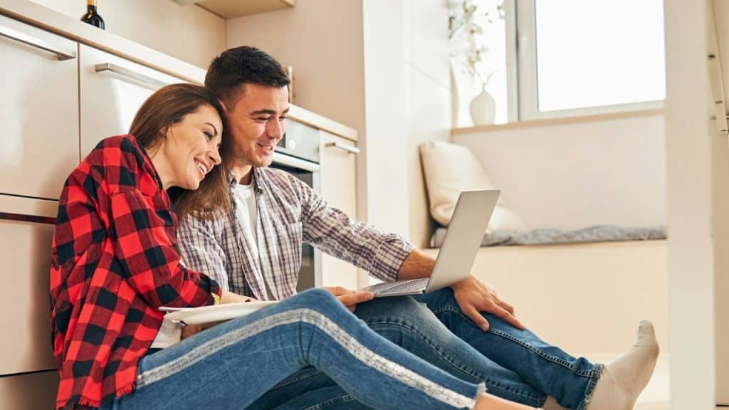 Smiling couple sitting on the kitchen floor, leaning against cabinets, and looking at a laptop.