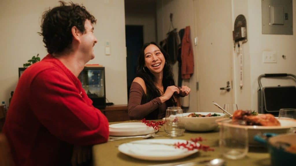 Man in a red sweater and a woman smiling and laughing at a dinner table in an apartment.