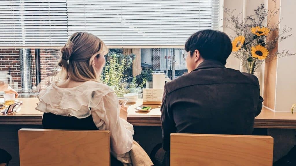 Two people sitting side-by-side facing a large window with blinds in a cafe.