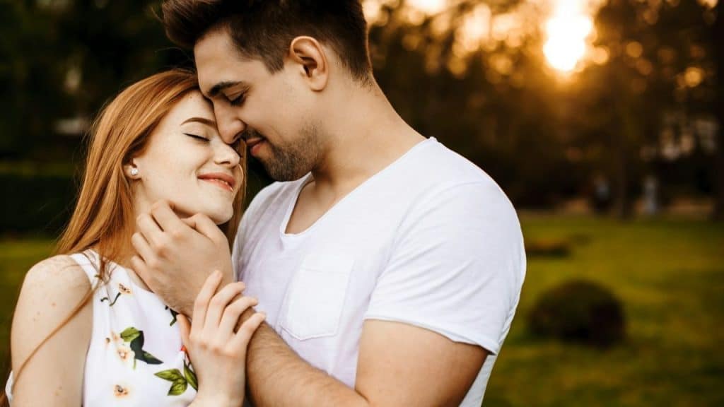 Close-up of a smiling couple embracing outdoors with the sun setting behind them.