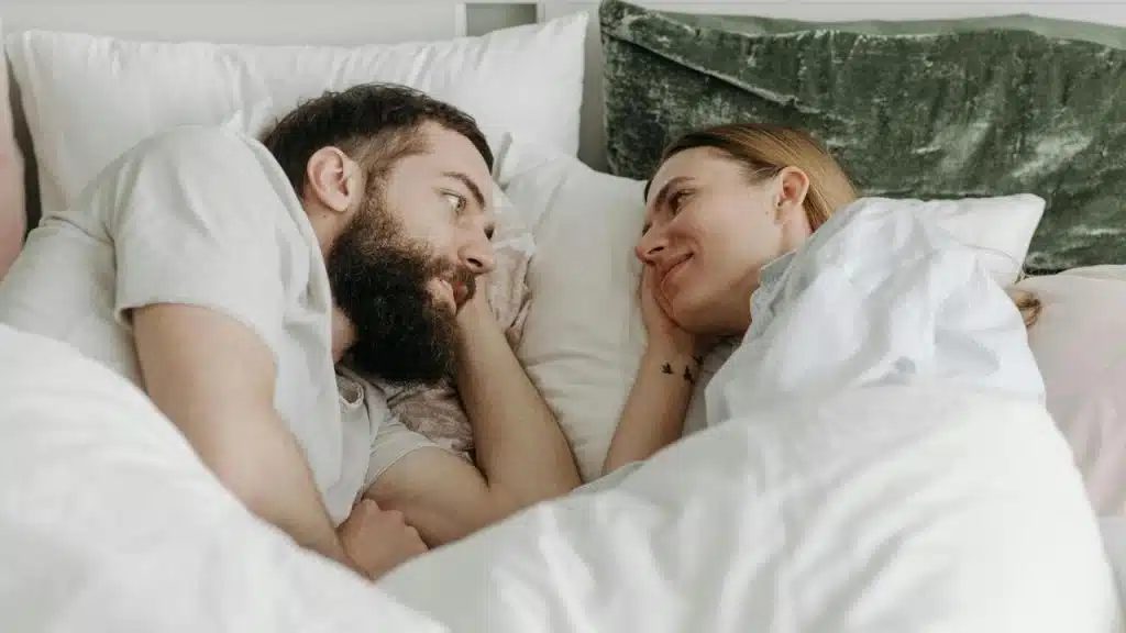 A couple lying in bed facing each other and smiling warmly.