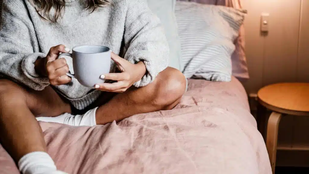 A person sitting on a bed holding a mug with both hands.