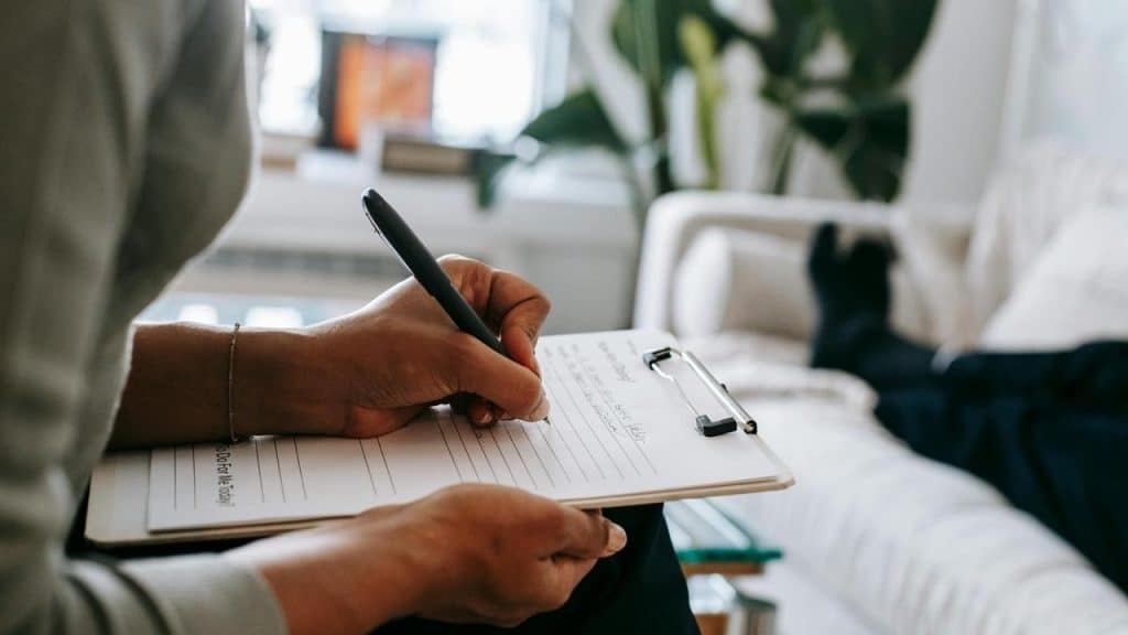 A person writing on a clipboard while another person lies on a couch in the background.