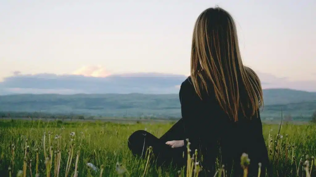 A woman sitting in a grassy field looking toward distant hills at sunset.