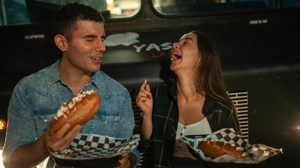 A man and woman sitting together eating hot dogs and laughing.