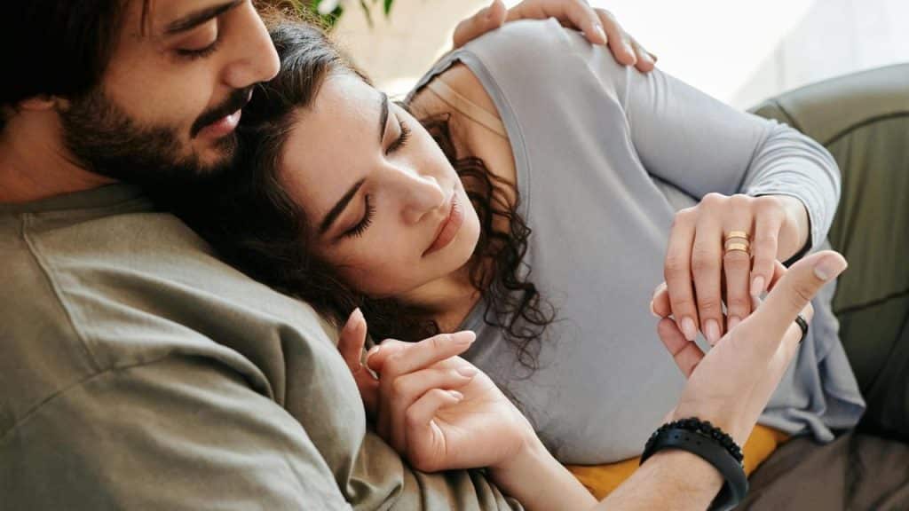 A couple cuddling on a couch while the woman gently touches the man’s hand.