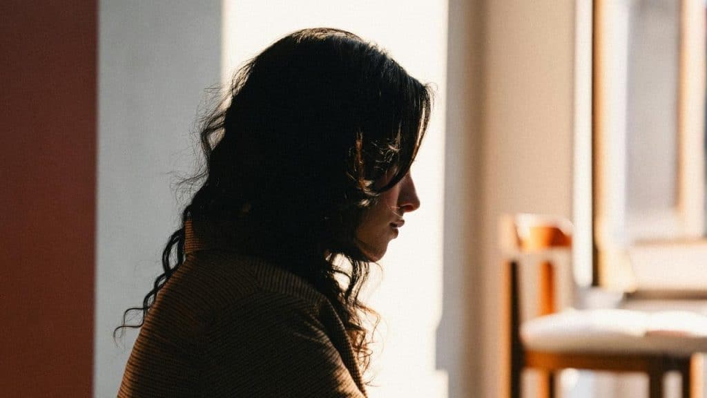 A woman sitting indoors in soft light, appearing deep in thought.