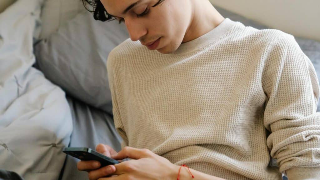A young man sitting on a bed while looking at his phone.