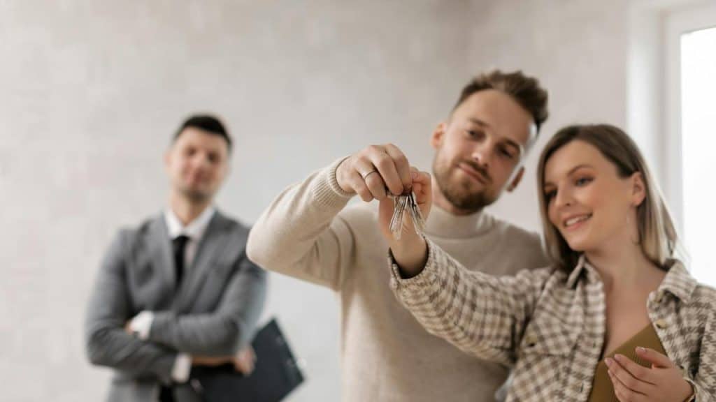 A smiling couple holding new house keys with a realtor standing behind them.