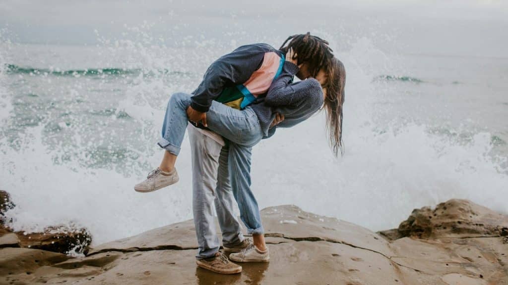 A man holding and kissing a woman as they stand on a rocky shore with a large wave crashing behind them.