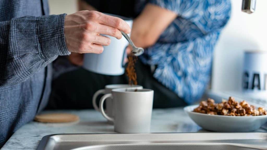 A person spooning coffee into mugs on a kitchen counter.