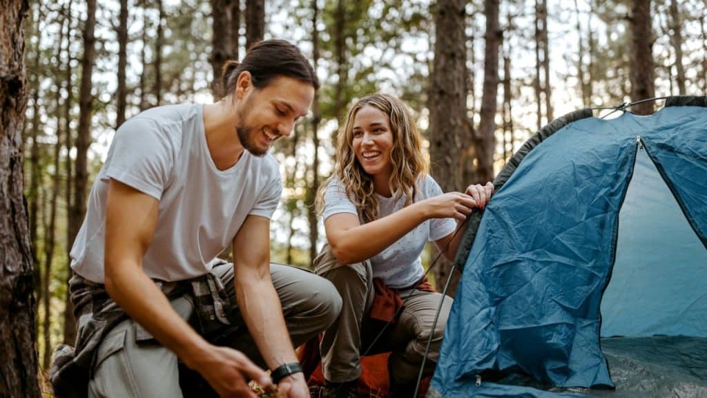 A smiling man and woman are kneeling in a forest, setting up a blue tent.