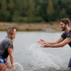 A man and woman laughing and splashing water at each other in a lake.