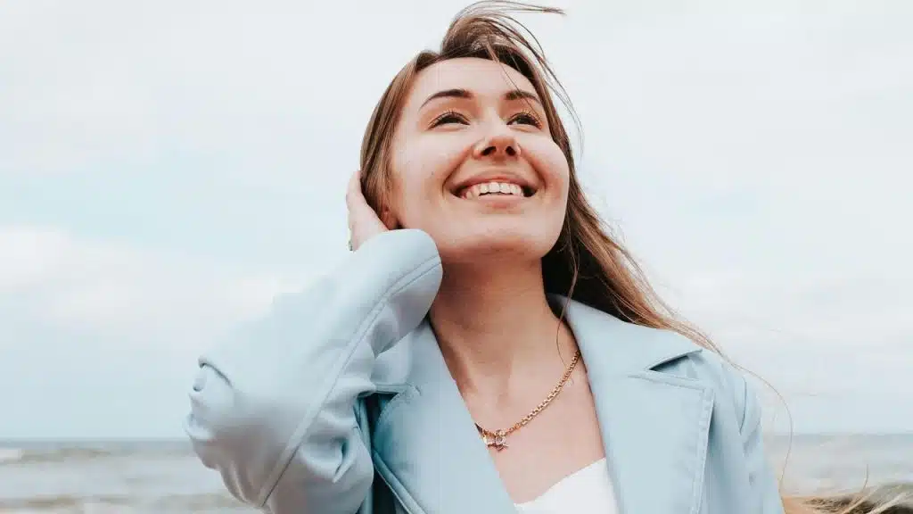 A woman smiling and looking up while standing by the beach on a windy day.