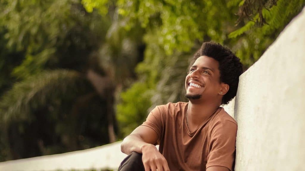 A man sitting outdoors smiling and looking up with greenery in the background.