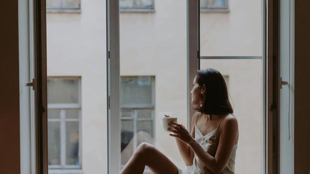A woman sitting on a windowsill holding a cup and looking outside.