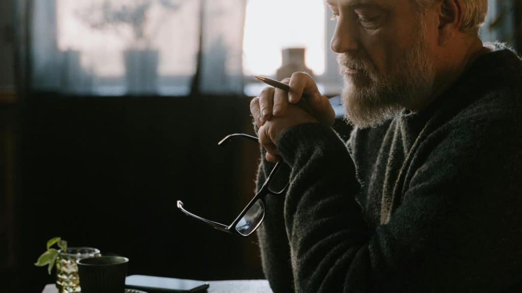 An older man sitting at a table holding glasses and a pen while thinking.